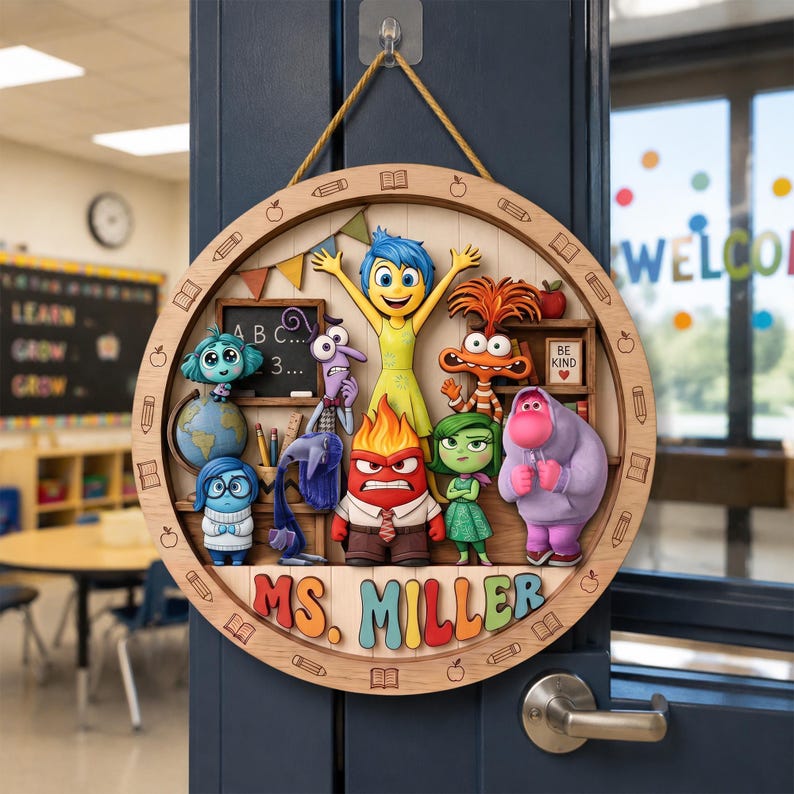 May include: A circular wooden classroom door sign featuring colorful 3D-style characters from Inside Out, including Joy, Sadness, Anger, Disgust, Fear, Bing Bong, and Anxiety. The sign displays the text MS. MILLER and is suspended by a gold rope.
