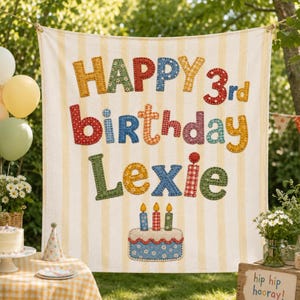 May include: A cream and yellow striped fabric banner featuring colorful, patterned fabric letters that spell out HAPPY 3rd birthday Lexie, with a stitched fabric birthday cake illustration at the bottom featuring three lit candles.