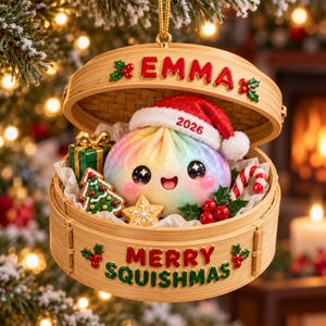 Peut inclure: Une décoration en bambou en forme de panier vapeur contenant une boulette arc-en-ciel portant un bonnet de Père Noël rouge avec 2026. Le couvercle indique EMMA et la base MERRY SQUISHMAS. On y voit un cadeau vert, des biscuits et un sucre d'orge.
