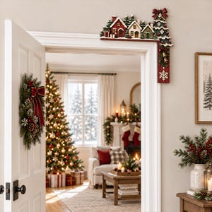 May include: A decorative wooden door corner topper featuring a miniature snowy village with red and white houses, pine trees, and a tall Christmas tree adorned with a red bow, gold bells, and a white snowflake accent on the vertical panel.