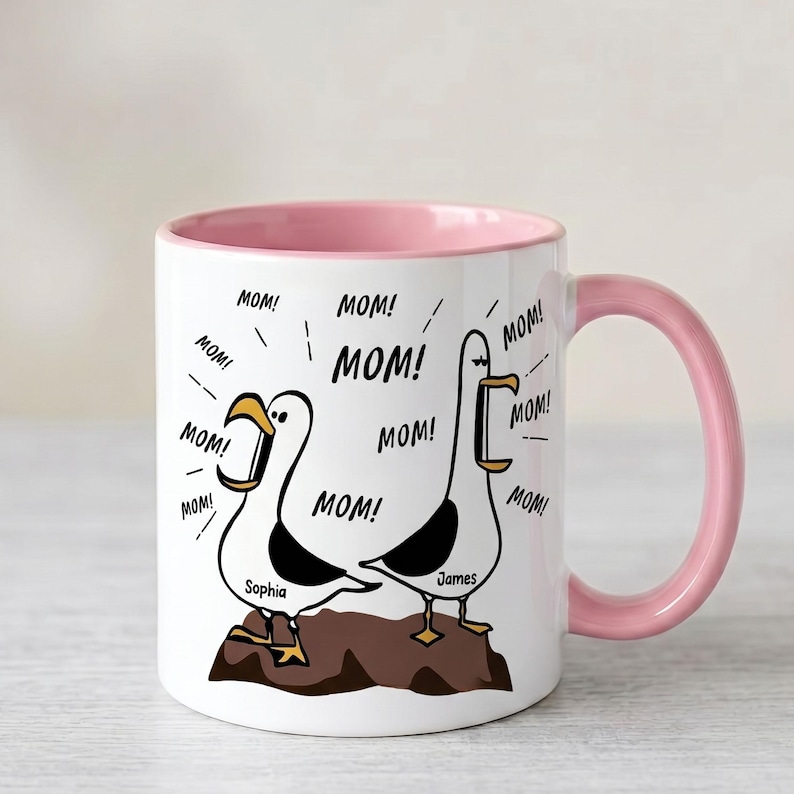 Peut inclure: Mug en c&eacute;ramique blanche avec int&eacute;rieur et anse roses. L'illustration repr&eacute;sente deux mouettes criant "MOM!". Les noms "Sophia" et "James" sont en dessous. Un cadeau amusant pour la f&ecirc;te des m&egrave;res.