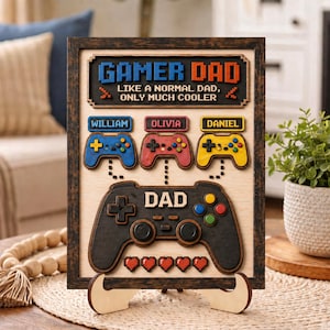 May include: A rectangular wooden wall sign in a dark frame featuring the text GAMER DAD LIKE A NORMAL DAD, ONLY MUCH COOLER. It includes three smaller game controllers labeled William, Olivia, and Daniel, and one large controller labeled DAD.