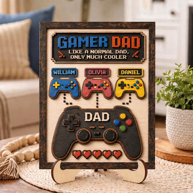 May include: A rectangular wooden wall sign in a dark frame featuring the text GAMER DAD LIKE A NORMAL DAD, ONLY MUCH COOLER. It includes three smaller game controllers labeled William, Olivia, and Daniel, and one large controller labeled DAD.