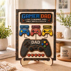 May include: A framed wooden sign featuring the text GAMER DAD LIKE A NORMAL DAD, ONLY MUCH COOLER. It includes three small game controllers labeled WILLIAM, OLIVIA, and DANIEL above a large black controller labeled DAD with five pixelated hearts.