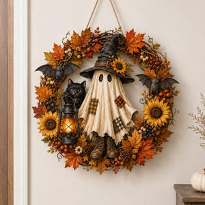 May include: A decorative wreath featuring a central ghost figure wearing a black witch hat and boots, accompanied by a black cat holding a lantern. The wreath is adorned with orange and brown autumn leaves, sunflowers, berries, and two bats.