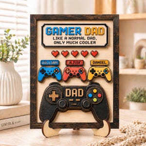 May include: A rectangular wooden wall sign with a dark frame featuring the text GAMER DAD LIKE A NORMAL DAD, ONLY MUCH COOLER. It includes five red pixelated hearts, three small game controllers labeled William, Olivia, and Daniel, and one large black controller labeled DAD.