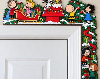May include: A Peanuts-themed Christmas door corner decoration featuring Linus, Sally, Snoopy, Woodstock, Charlie Brown, Lucy, Marcie, and Peppermint Patty. The colorful wooden cutout displays characters with gifts, a red doghouse, and a decorated tree.