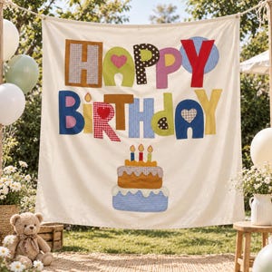 May include: A cream-colored fabric banner featuring the words HAPPY BIRTHDAY in colorful, patterned fabric letters. Below the text is a two-tiered cake applique with three lit candles. The banner is suspended outdoors with balloons.