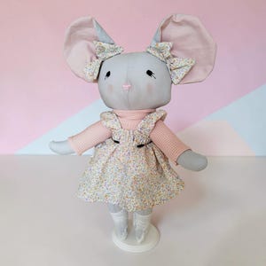 Puede incluir: Una muñeca de ratón gris hecha a mano con grandes orejas rosas y un lazo floral. La muñeca lleva una camisa rosa de manga larga y un vestido con estampado floral. Se apoya sobre una base blanca y tiene calcetines blancos y zapatillas de ballet.