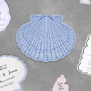 May include: A collection of wedding stationery featuring shell-shaped cards in blue and white, including a menu, RSVP card with a QR code, details card, and a save the date. Two white daisy flowers and two small seashells are arranged among the cards.
