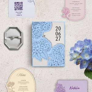 May include: A wedding stationery set featuring a light blue invitation with hydrangea illustrations and the date 20 06 27, an RSVP card with a QR code, a details card, a menu, a place card labeled Natalia, and a ring in an octagonal box.