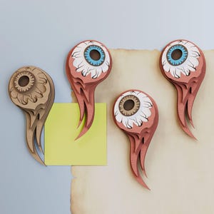 May include: Four layered paper cutouts shaped like stylized eyes with trailing tendrils. Two eyes feature blue irises, one has a brown iris, and one is monochromatic tan. A bright yellow square note is positioned behind the tan eye cutout.