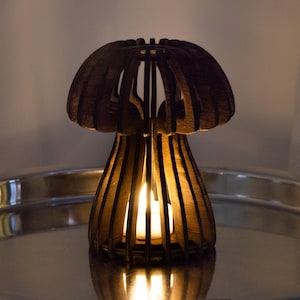 Op de afbeelding: Een houten kandelaar in de vorm van een paddenstoel. Het donkerbruine, lasergesneden ontwerp heeft een kap en een uitlopende basis, met verticale lamellen die een open structuur creëren. Een brandende kaars binnenin geeft een warme gloed.