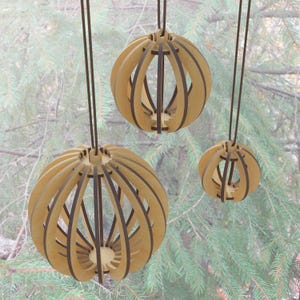 Puede incluir: Tres adornos esféricos de madera colgantes con acabado de madera natural y detalles en marrón oscuro. Las formas esféricas están construidas con listones de madera curvados, creando un diseño abierto. Cada adorno cuelga de un fino cordón marrón oscuro.