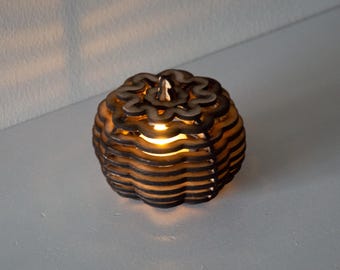 May include: A decorative wooden candle holder shaped like a pumpkin. The pumpkin is made of layered, laser-cut wood with a warm light emanating from within. The design features a scalloped top and a round body.