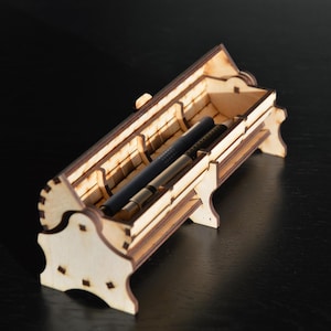 Può includere: Un portapenne in legno chiaro di forma rettangolare con intagli decorativi. Il portapenne contiene due penne, una nera e una dorata. Il portapenne è progettato con un coperchio e supporti laterali ed è posto su una superficie scura.