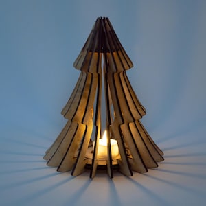 Puede incluir: Un portavelas de madera con forma de árbol de Navidad. El portavelas está hecho de madera cortada con láser en capas, con una vela de té en el interior. La cálida luz de la vela proyecta sombras. El diseño del árbol es una decoración festiva.