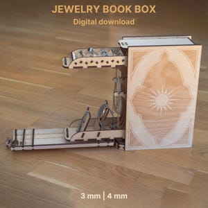 May include: A wooden jewelry book box with the text "JEWELRY BOOK BOX" and "Digital download". The box is open, revealing compartments for rings, necklaces, and other jewelry. The design includes a sunburst pattern and is made of light-colored wood.