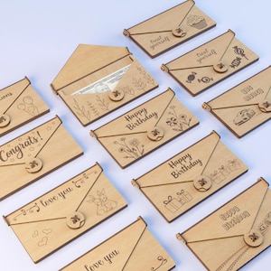 Puede incluir: Una colección de tarjeteros de madera con varios diseños y textos grabados. Algunos tarjeteros dicen "¡Felicidades!", "Feliz Cumpleaños" y "Te amo". Otros presentan imágenes de una magdalena, caramelos y un coche. Cada uno tiene un cierre de botón.