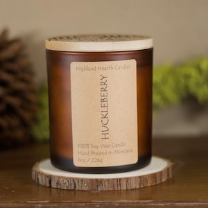 May include: A brown glass candle jar with a wooden lid and a label that reads "HUCKLEBERRY." The candle sits on a small wooden coaster. The label also states "100% Soy Wax Candle Hand Poured in Montana 8oz / 226g."