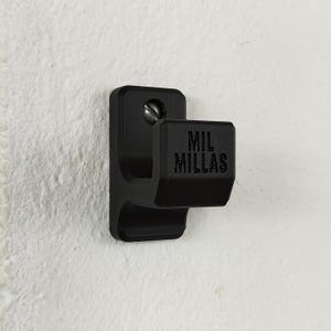 Pode incluir: Um gancho de parede preto com o texto "MIL MILLAS" está montado numa parede branca. O gancho é feito de um material sólido e é fixado com um parafuso. O design é simples e funcional.