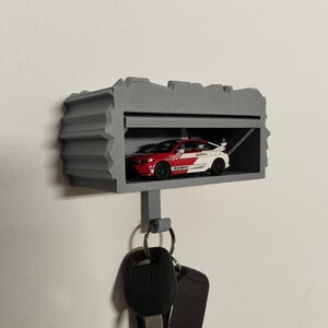 Könnte beinhalten: Ein grauer, 3D-gedruckter Schlüsselhalter in Form einer Garage mit einem rot-weißen Spielzeugauto im Inneren. Der Schlüsselhalter hat einen Haken für Schlüssel und ist an einer weißen Wand befestigt.
