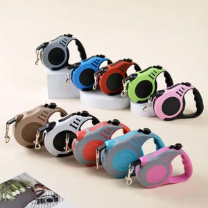 May include: A selection of retractable dog leads in a range of colours: grey, blue, red, green, brown, white, and pink. Each lead has a black handle and a chrome clasp for attaching to a dog's collar.