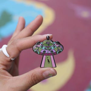 May include: A colorful UFO-shaped enamel pin with a detailed design in shades of green, purple, and pink. The pin features a small alien figure and a brain graphic. The pin is held up by a hand.