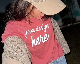 May include: A dusty rose t-shirt with the text "your design here" in white script. The person is wearing a tan trucker hat and a beige and grey jacket. The person is also wearing blue jeans.