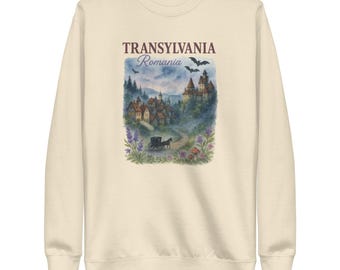 Puede incluir: Sudadera color crema con una ilustración en acuarela de un pueblo europeo. La obra de arte representa un castillo, casas, un carruaje tirado por caballos y murciélagos, con el texto "TRANSYLVANIA Romania".