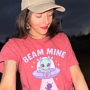 Könnte beinhalten: Rosafarbenes T-Shirt mit dem Text "BEAM MINE" und einem Cartoon-Alien in einem UFO. Das Alien winkt. Die Person trägt eine beige Baseballkappe mit dem Wort "Grateful" auf der Vorderseite.