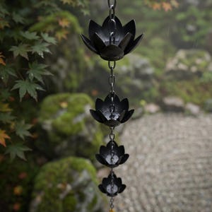 May include: A black metal rain chain with four lotus flower-shaped cups suspended from a chain. The rain chain hangs in front of a blurred background of green foliage and a stone garden.