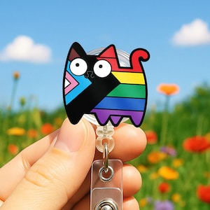 May include: A cat-shaped retractable badge reel with a colorful design. The cat features a black body, rainbow stripes, and a pink, blue, and black triangle. The reel has a clear plastic clip and a metal button.