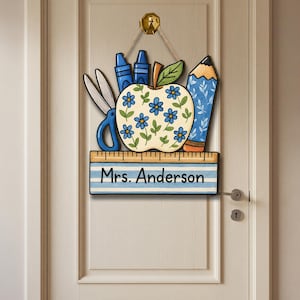 May include: A wooden teacher's door sign featuring an apple decorated with blue flowers, scissors, crayons, and a pencil. The sign includes the name "Mrs. Anderson" on a ruler-themed banner. The sign is hanging on a white door.