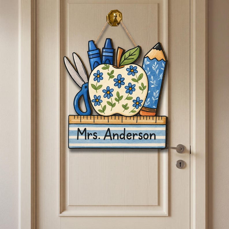 May include: A wooden teacher's door sign featuring an apple decorated with blue flowers, scissors, crayons, and a pencil. The sign includes the name "Mrs. Anderson" on a ruler-themed banner. The sign is hanging on a white door.