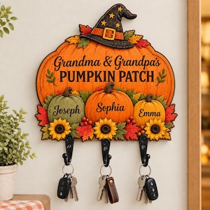 Może przedstawiać: Dekoracyjny ścienny wieszak na klucze w kształcie dużej pomarańczowej dyni z czarnym kapeluszem czarownicy. Napis głosi Grandma & Grandpa's PUMPKIN PATCH. Trzy mniejsze dynie są podpisane Joseph, Sophia i Emma, z trzema czarnymi haczykami.
