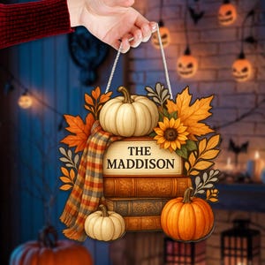 Könnte beinhalten: Holzschild im Herbststil mit Kürbissen, Sonnenblumen und Blättern. Das Schild, geformt wie Bücher, trägt den Text "THE MADDISON". Ein karierter Schal liegt über den Büchern. Das Schild wird von einer weißen Schnur gehalten.
