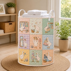 Op de afbeelding: Een cilindrische stoffen wasmand met een doorgestikte textuur en een rasterpatroon van negen illustraties met witte ganzen die verschillende dagelijkse activiteiten uitvoeren. De mand heeft twee witte stoffen handvatten en bevat opgevouwen witte handdoeken.