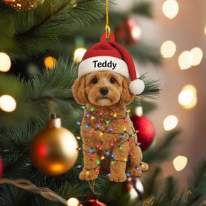 May include: A Christmas tree ornament featuring a brown dog wearing a Santa hat with the name "Teddy" and a string of colorful lights. The ornament is hanging on a Christmas tree with gold and red ornaments.
