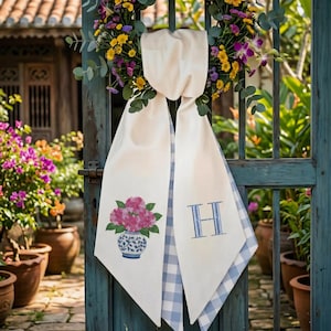 May include: Cream-colored decorative scarf hanging on a teal gate, adorned with a floral wreath. One side features a pink floral arrangement in a blue and white vase, the other a blue embroidered "H" over a blue and white gingham pattern.