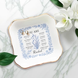 May include: White ceramic trinket dish with a gold rim, featuring blue floral designs and the words "YOU ARE" with inspirational affirmations. Two pearl earrings rest on the dish. A thoughtful gift.