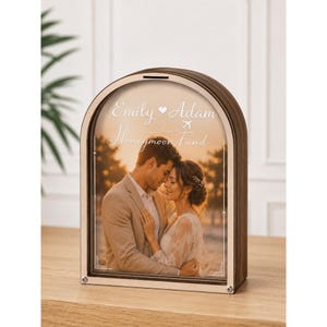 May include: An arched wooden shadow box money bank with a clear front panel. The insert features a photograph of a couple embracing, with the printed text "Emily Adam" separated by a heart, a small airplane icon, and the words "Honeymoon Fund".