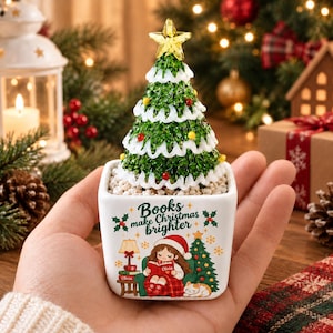 May include: A small white square planter featuring a green Christmas tree with a yellow star topper. The planter displays the text "Books make Christmas brighter" above an illustration of a person reading a book with a cat and a small tree.