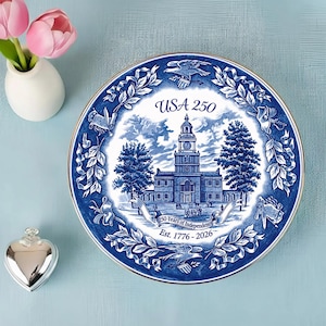 May include: A blue and white decorative plate with a gold rim, featuring a detailed illustration of a building with a clock tower, trees, and the text "USA 250". The plate commemorates 250 years of independence.