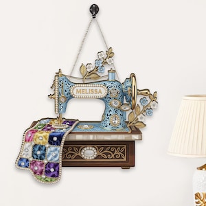 Op de afbeelding: Decoratieve wanddecoratie in de vorm van een vintage naaimachine. De lichtblauwe machine heeft gouden accenten en de naam "Melissa". Een kleurrijke quilt is over de basis gedrapeerd. Bloemen en edelstenen completeren het ontwerp.