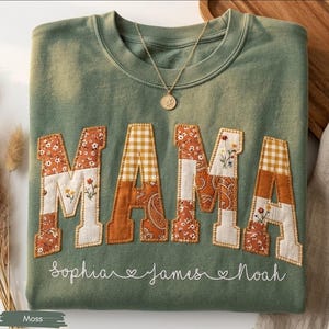 May include: A moss green sweatshirt with the word "MAMA" appliquéd in patchwork fabric. The letters feature floral, paisley, and gingham patterns. Below the word are the names "Sophia", "James", and "Noah". A gold necklace with a pendant rests on the shirt.
