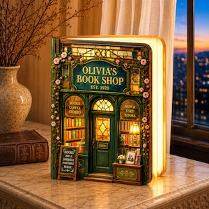 May include: A book-shaped diorama featuring a miniature bookstore titled Olivia's Book Shop Est. 1928. The green facade is adorned with pink flowers, illuminated windows, and a sign reading Books Are A Uniquely Portable Magic. The interior displays shelves of books and a lamp.