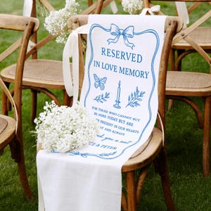 Op de afbeelding: Een witte stoffen stoelversiering met blauwe tekst en illustraties, waaronder een strik, vlinder, kaars en bladeren. De tekst luidt "RESERVED IN LOVE MEMORY" en een toewijding. De versiering is over een houten stoel gedrapeerd.
