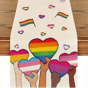 Puede incluir: Camino de mesa color crema con un diseño de corazones y banderas arcoíris, y manos sosteniendo corazones con varios diseños de banderas del orgullo LGBTQ+. El camino de mesa está decorado con corazones y banderas de varios colores.
