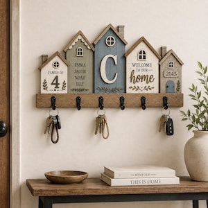 Op de afbeelding: Een houten wand-sleutelhouder met vijf huisvormige panelen in blauw, groen en wit. De panelen bevatten de tekst "FAMILY OF 4", namen, een grote letter "C", "WELCOME TO OUR home" en "2145". Voorzien van zes zwarte metalen haken voor sleutels.